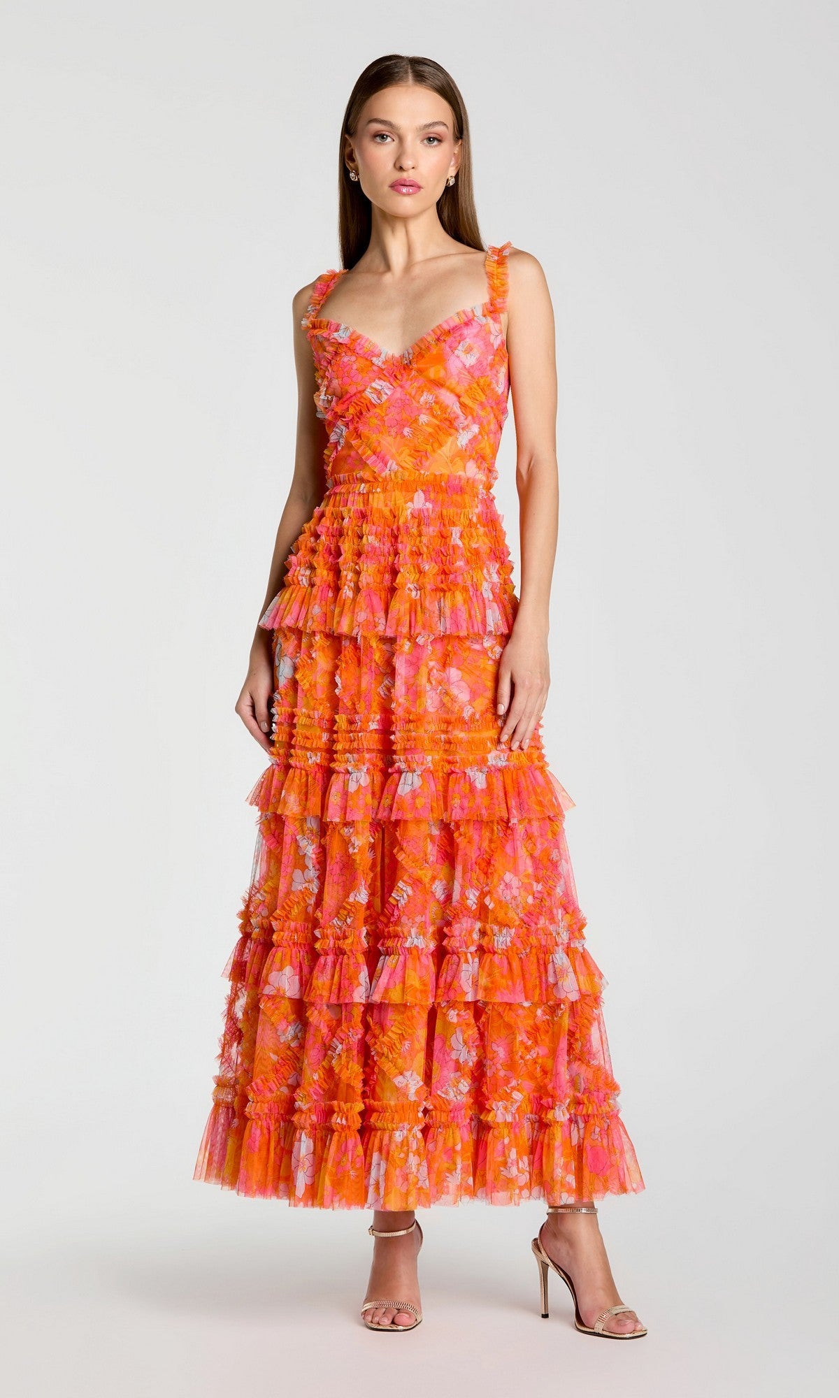 A woman stands in neutral heels against a plain light background, wearing the Mac Duggal 8475 Long Formal Dress in Orange Multi—a sleeveless, floor-length fit-and-flare gown featuring tiered ruffles and a vibrant floral pattern in orange and pink.