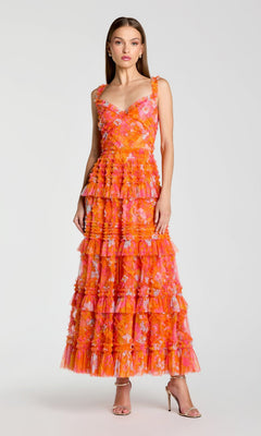 A woman stands in neutral heels against a plain light background, wearing the Mac Duggal 8475 Long Formal Dress in Orange Multi—a sleeveless, floor-length fit-and-flare gown featuring tiered ruffles and a vibrant floral pattern in orange and pink.