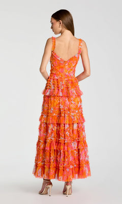 A woman stands facing away, wearing the Mac Duggal 8475 long formal dress— a sleeveless, floor-length, layered fit-and-flare gown in orange and pink with floral details and ruffles—paired with beige high-heeled sandals.