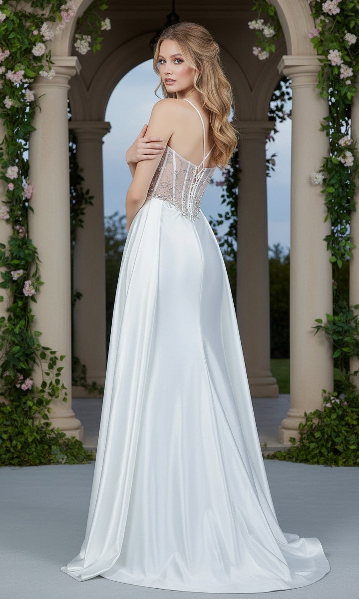 A woman in the Cinderella Couture 8483J long prom dress, featuring a white sleeveless design with a sheer corset bodice and intricate beaded embroidery, stands outdoors before columns and greenery, looking over her shoulder.