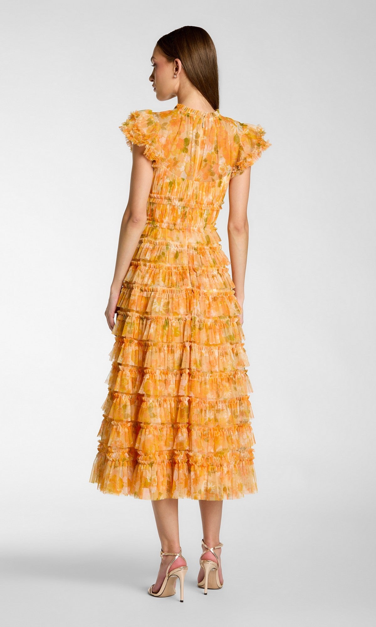 A woman stands facing away, modeling the Mac Duggal 8497 Tea-Length Party Dress in orange floral, featuring tiered ruffles and short sleeves, paired with high-heeled sandals against a plain gray background.