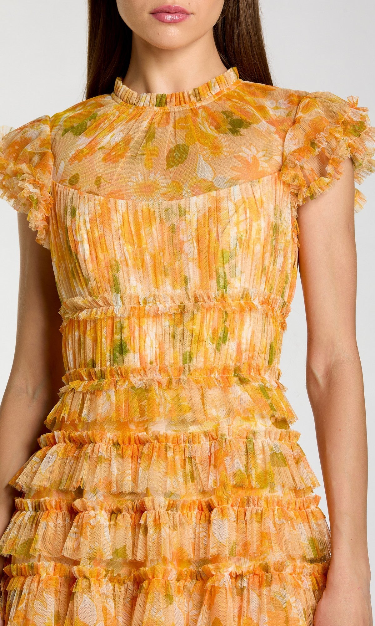 A woman wearing the Tea-Length Party Dress: Mac Duggal 8497, a short-sleeve pleated tea-length dress featuring ruffles, a yellow, orange, and green floral pattern, a high neckline, and a sheer upper bodice.
