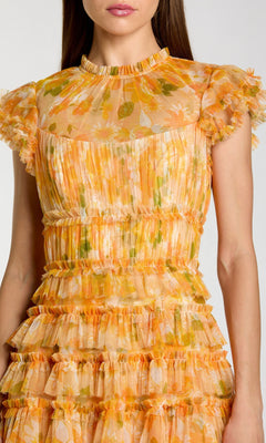 A woman wearing the Tea-Length Party Dress: Mac Duggal 8497, a short-sleeve pleated tea-length dress featuring ruffles, a yellow, orange, and green floral pattern, a high neckline, and a sheer upper bodice.