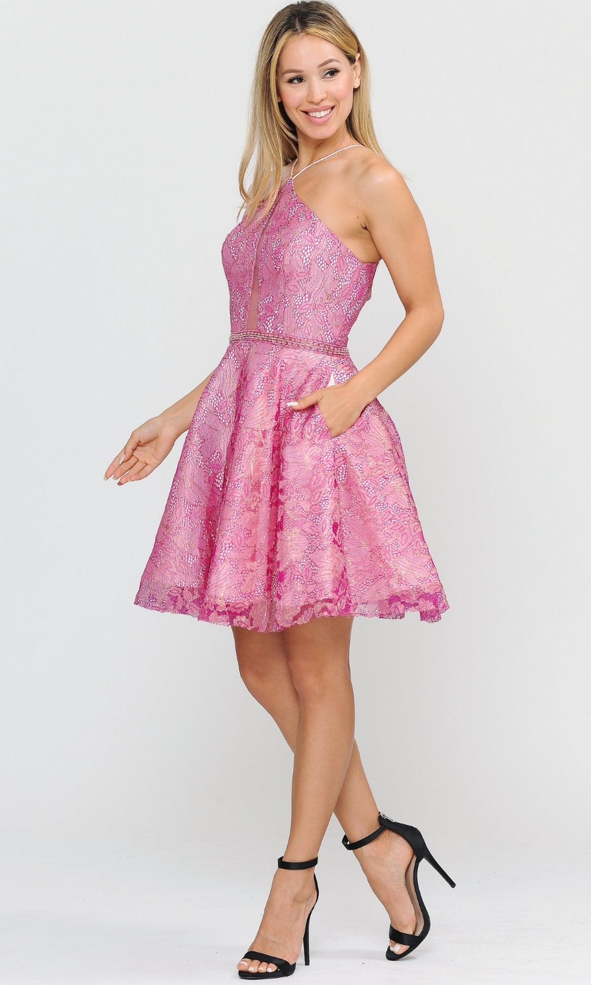 A woman wearing the Glitter-Mesh Short A-Line Homecoming Dress 8506, a sleeveless pink lace dress with side pockets, paired with black high-heeled sandals, stands against a plain light background.
