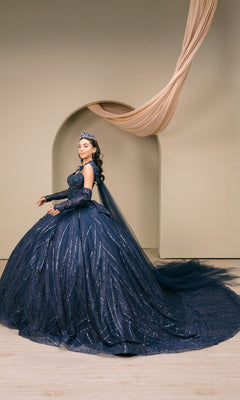 A woman wearing the Quinceanera Dress 8510JQ by Cinderella Couture—a dark blue, sequin-embellished ball gown—and a crown stands before an arched backdrop draped with fabric, radiating timeless elegance.