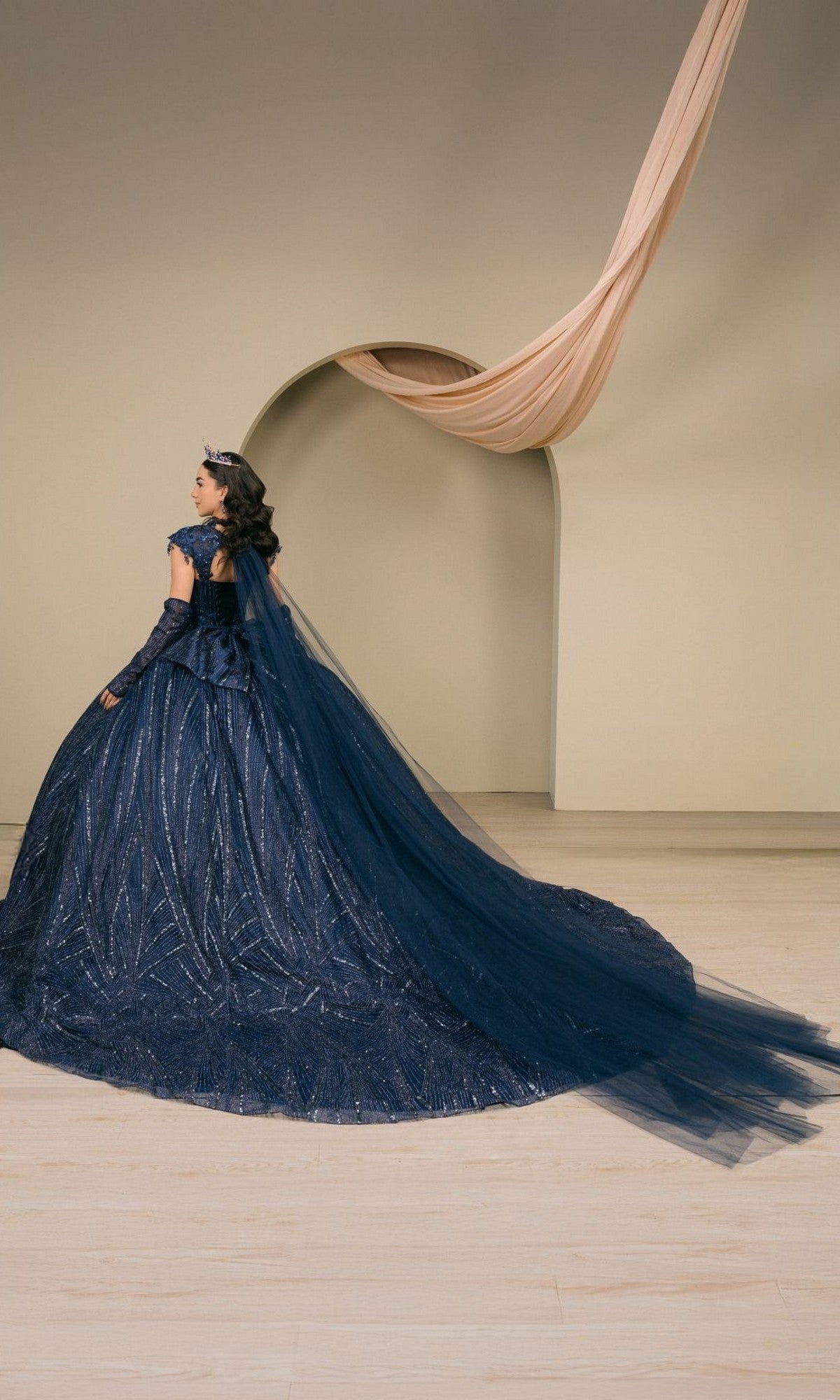 A woman in a navy blue quinceañera ball gown with a long train and crown stands in front of a beige wall with an arch and draped fabric. The stunning Quinceanera Dress 8510JQ by Cinderella Couture features elegant sequin-embellished details, adding a touch of sparkle to her regal look.