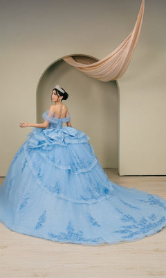 A person wearing the Quinceanera Dress 8520JQ By Cinderella Couture—an ornate light blue ball gown featuring a sparkling corset bodice and a tiara—stands in front of a beige arched doorway with fabric elegantly draped overhead.