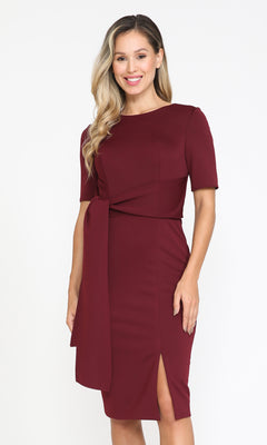 A woman stands against a white background wearing the Short Sleeve Classy Knee-Length Party Dress 8524 in burgundy, featuring short sleeves, a side knot detail, and a front slit—an elegant choice for semi-formal or work occasions.
