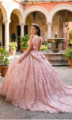 A young woman wearing the Quinceanera Dress 8530JQ by Cinderella Couture, featuring a beaded corset bodice and a sparkling tiara, stands in a courtyard near a stone fountain and potted plants, looking over her shoulder.