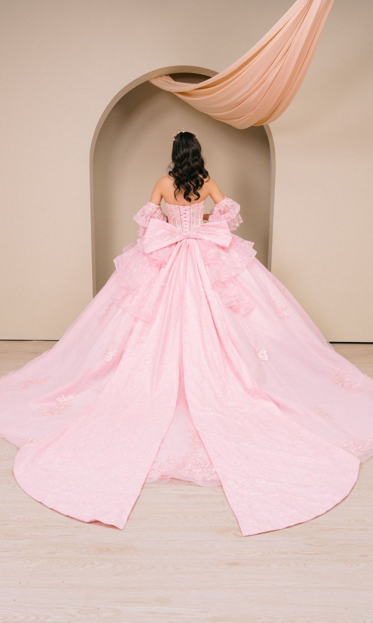 A person stands facing an arched backdrop with draped fabric overhead, wearing the Quinceanera Dress 8535JQ by Cinderella Couture—a light pink, off-the-shoulder ball gown featuring a large bow and long train.