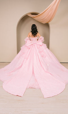 A person stands facing an arched backdrop with draped fabric overhead, wearing the Quinceanera Dress 8535JQ by Cinderella Couture—a light pink, off-the-shoulder ball gown featuring a large bow and long train.