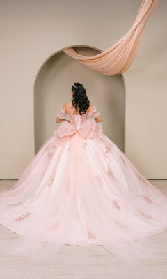 A woman wearing the Quinceanera Dress 8540JQ by Cinderella Couture, a blush pink gown with lace details, stands facing away in front of a beige arch adorned with draped fabric.
