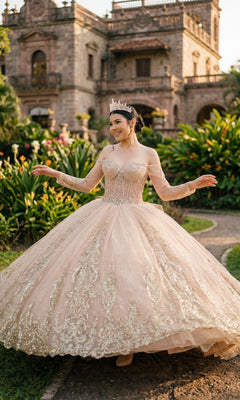 A woman in the Quinceanera Dress 8545JQ By Cinderella Couture, a pink ball gown with beaded sequin details and an off-shoulder sweetheart neckline, smiles outdoors on a garden path with a historic stone building behind her.