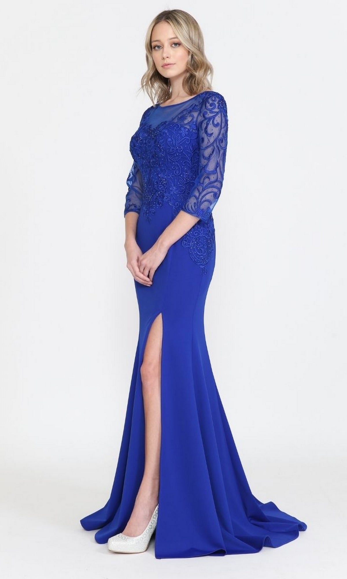 A woman stands against a plain light background wearing the Embroidered Sheer-Sleeved Long Prom Dress 8564 in royal blue, featuring lace detailing on the bodice and sleeves, a front slit, and paired with white high-heeled shoes.