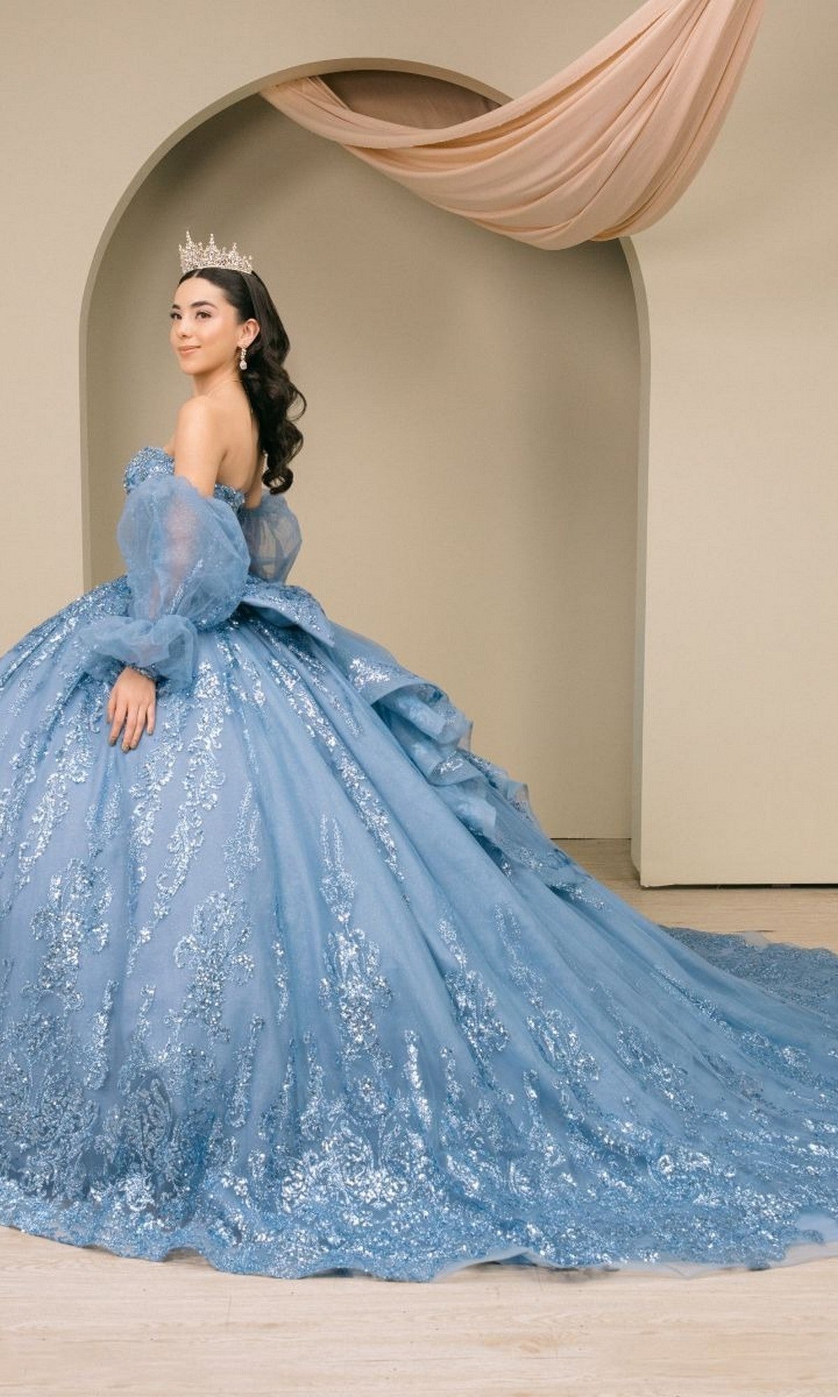 A woman poses in front of a beige archway with draped fabric overhead, wearing the Quinceanera Dress 8565JQ By Cinderella Couture—a light blue, off-shoulder sequin ball gown featuring an embellished corset bodice and a crown.
