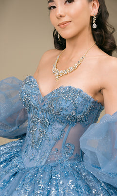 A person wears a blue, off-the-shoulder Cinderella Couture 8565JQ ballgown with a beaded, corset-style bodice and removable puff sleeves, accessorized with a crystal necklace and earrings.