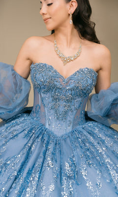 A woman photographed from the shoulders down wears the Quinceanera Dress 8565JQ By Cinderella Couture—a strapless, sparkly blue ball gown styled with removable puff sleeves and a gold necklace, evoking timeless Cinderella-inspired elegance.