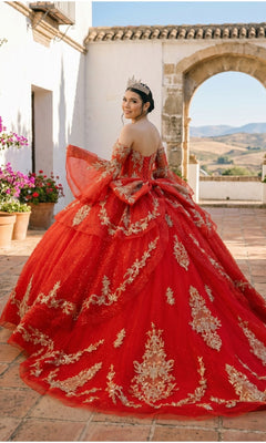A young woman in the Quinceanera Dress 8575JQ By Cinderella Couture, adorned with red and gold embellishments and a sparkling tiara, stands outdoors on a tiled patio facing slightly away, with hills and a stone archway visible in the background.