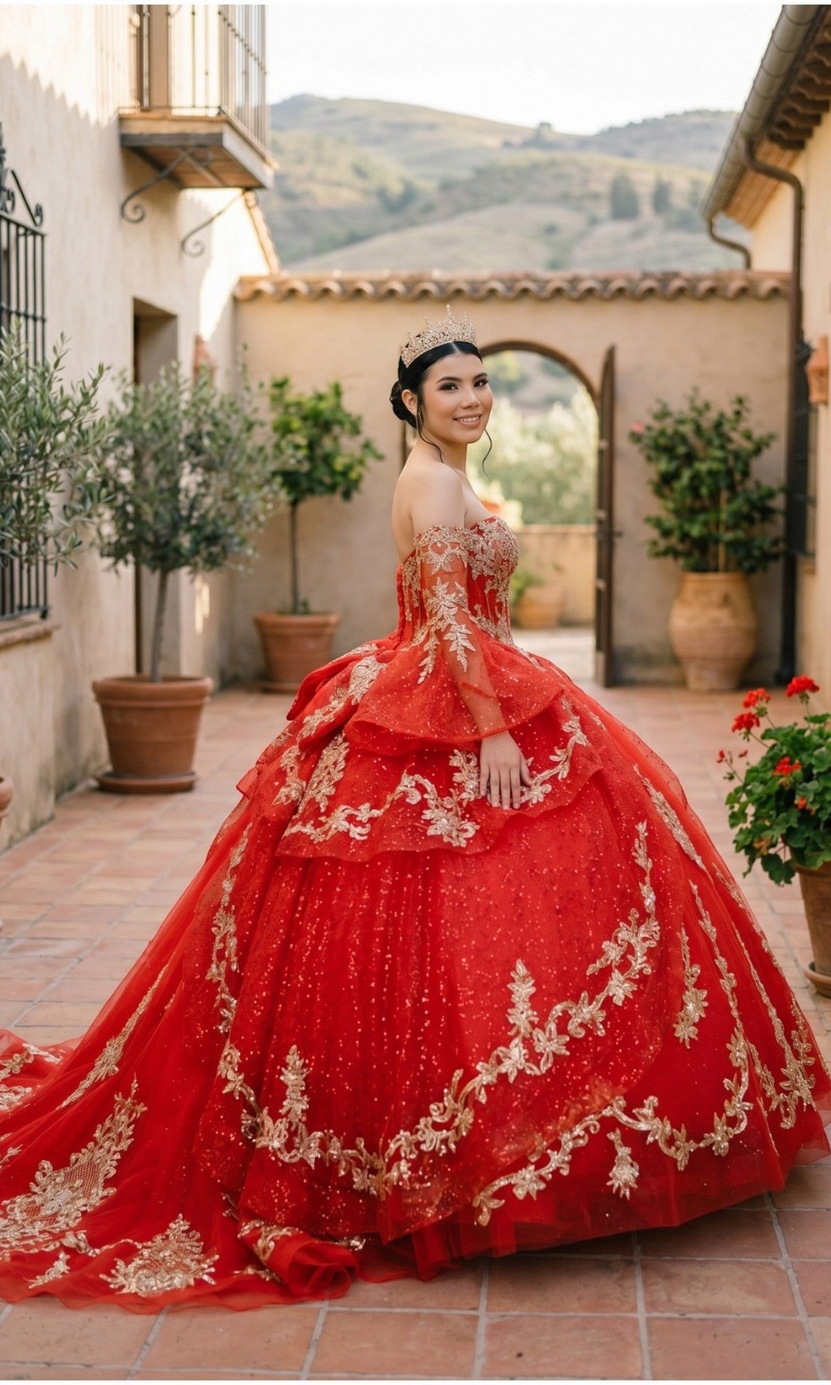 A young woman stands smiling in a courtyard adorned with potted plants and a scenic backdrop, radiating elegance in the Quinceanera Dress 8575JQ By Cinderella Couture—a glitter tulle embellished ball gown paired perfectly with her tiara, reminiscent of a stunning quinceañera celebration.