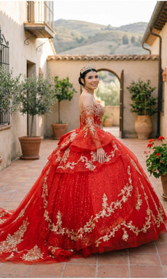 A young woman stands smiling in a courtyard adorned with potted plants and a scenic backdrop, radiating elegance in the Quinceanera Dress 8575JQ By Cinderella Couture—a glitter tulle embellished ball gown paired perfectly with her tiara, reminiscent of a stunning quinceañera celebration.