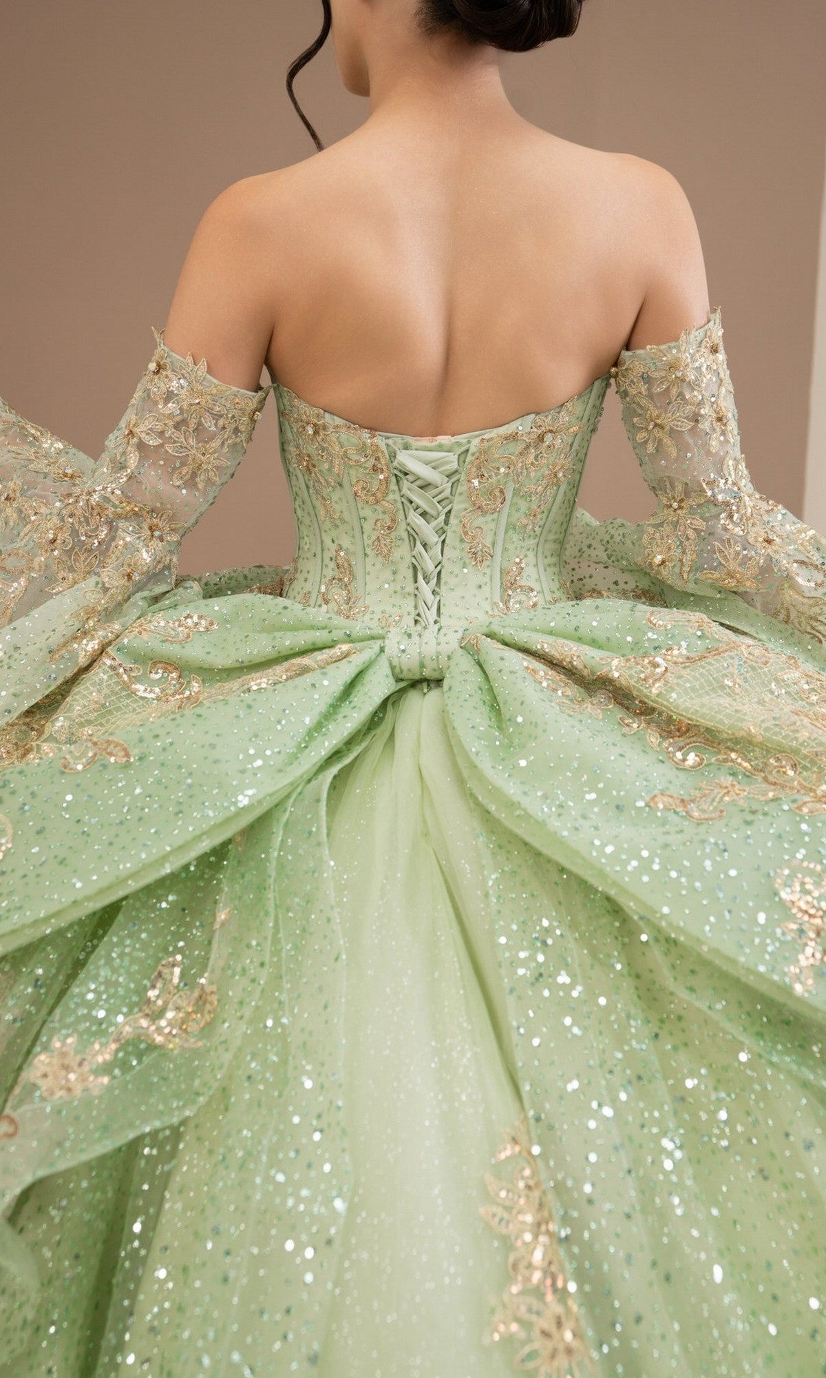 A woman wearing the Quinceanera Dress 8575JQ by Cinderella Couture—a sparkly, light green off-the-shoulder ball gown featuring gold embroidery, a lace-up back, and embellished details—photographed from behind.