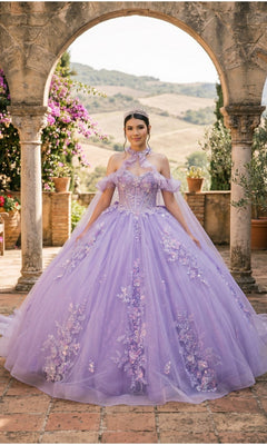 A young woman stands under a stone archway, wearing the Quinceanera Dress 8585JQ By Cinderella Couture in lilac, featuring a floral lace corset and delicate tiara, as the glitter tulle skirt shimmers while she poses outdoors with greenery and hills in the background.