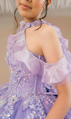 Wearing the Quinceanera Dress 8585JQ By Cinderella Couture, a lavender off-shoulder gown adorned with ruffled floral lace, appliqué, sequins, and a glitter tulle skirt, the person poses indoors.