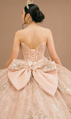 A woman is seen from behind against a neutral background, wearing the Quinceanera Dress 8590JQ By Cinderella Couture—a pink, glittery ball gown featuring a beaded corset bodice and a large bow at the back. She completes the look with a tiara.