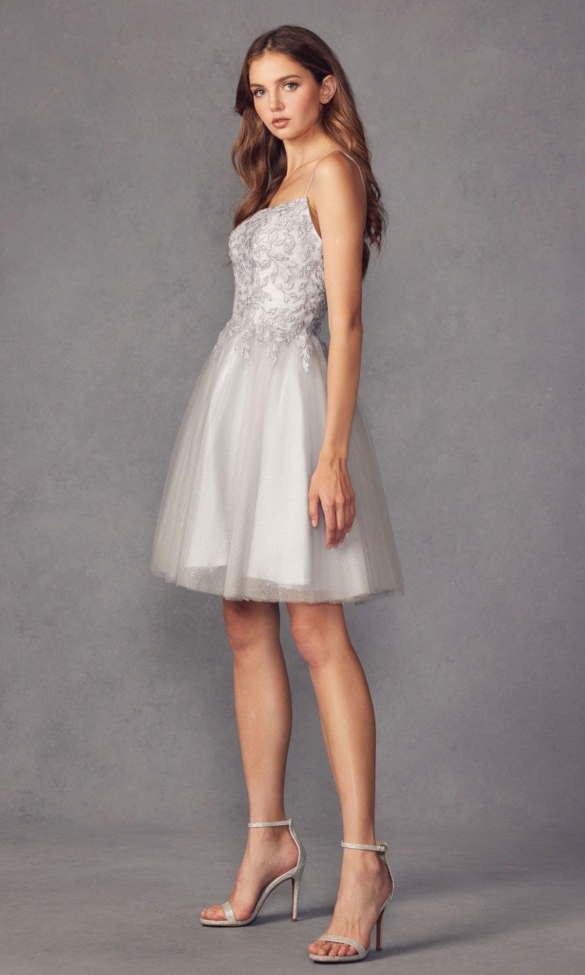 A woman in the Embroidered-Corset Babydoll Homecoming Dress 860, featuring silver floral embroidery and a tulle skirt, stands in profile against a gray background, wearing high-heeled sandals.