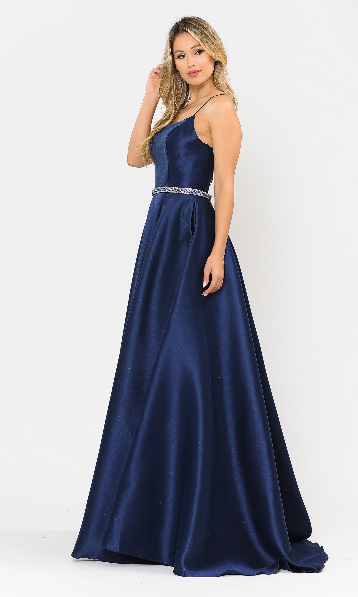 A woman poses against a plain white background wearing the Scoop-Neck Long A-Line Prom Dress with Pockets 8684—a navy blue, floor-length satin gown featuring spaghetti straps and a jeweled waist belt.