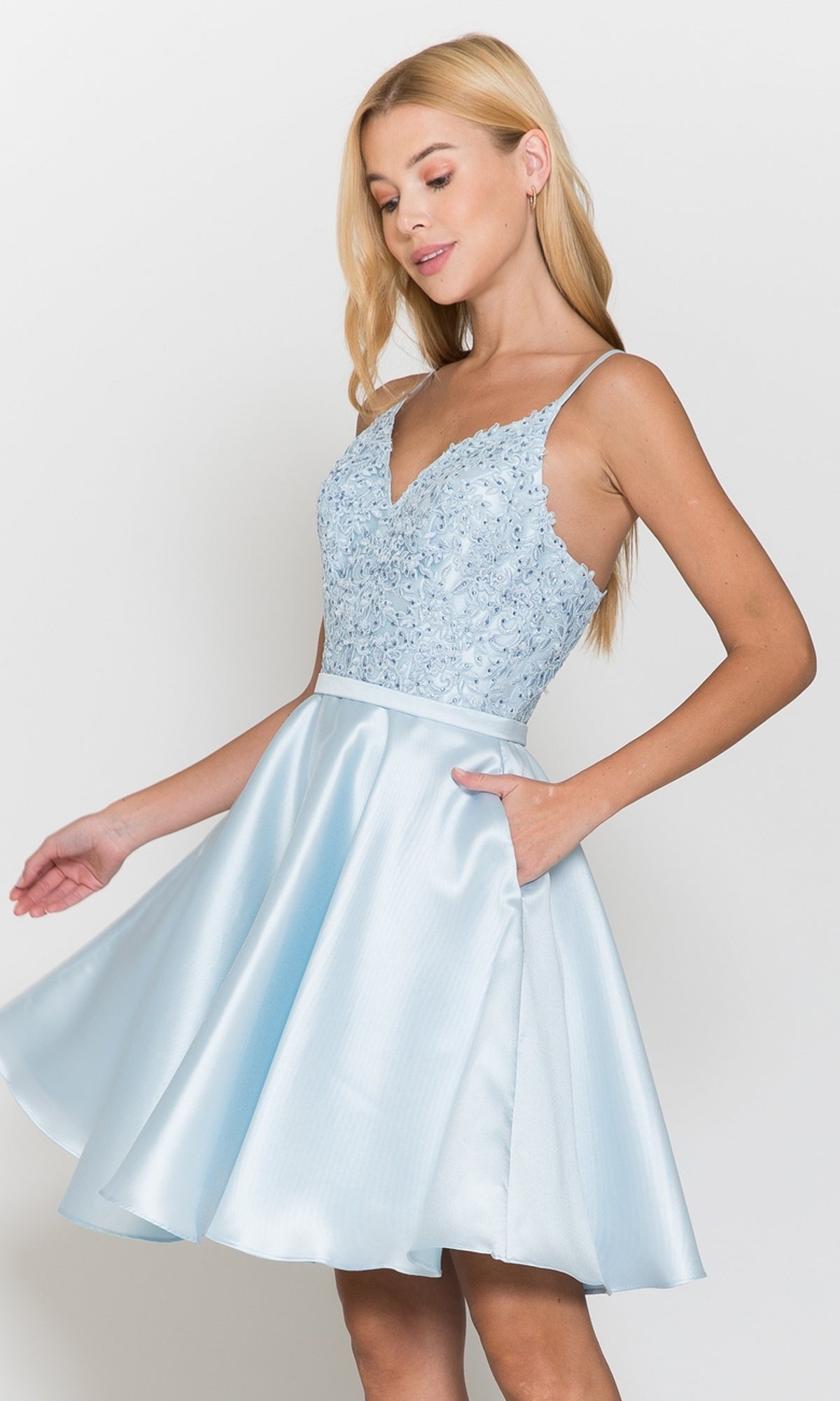 Woman modeling the Short A-Line Homecoming Dress with Pockets 8694 in blue, featuring a lace bodice and flared satin skirt, poses against a plain white background.
