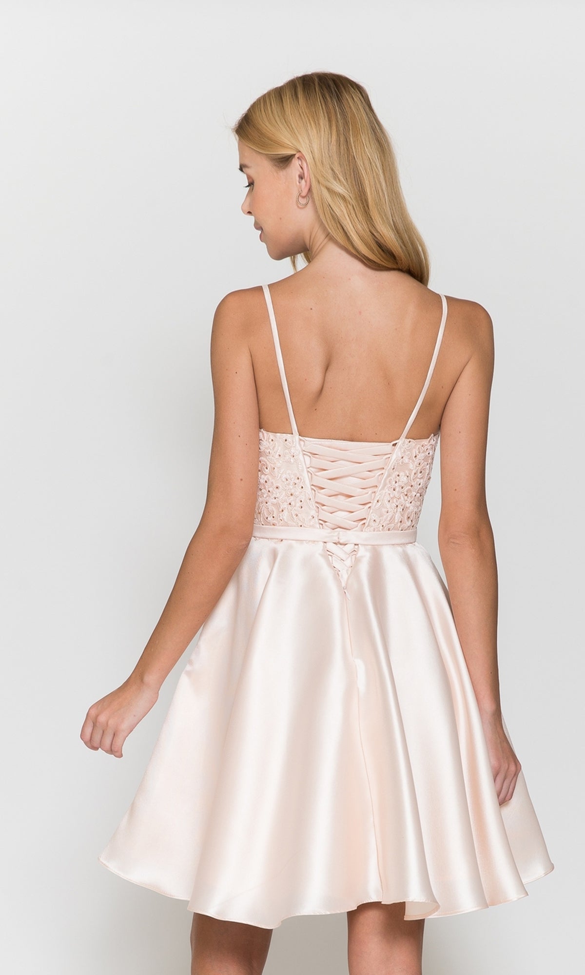 Woman in the Short A-Line Homecoming Dress with Pockets 8694—light pink satin, thin straps, lace-up back, and flared skirt—stands facing away from the camera.