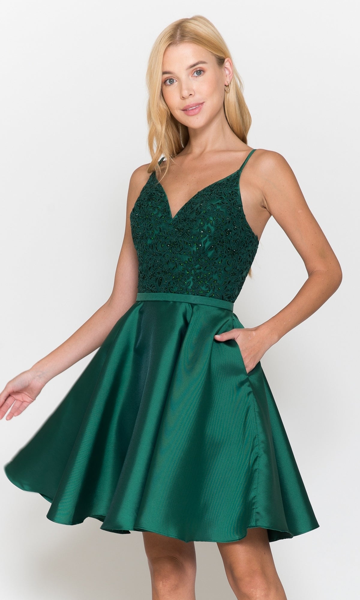 A woman stands against a plain white background wearing the Short A-Line Homecoming Dress with Pockets 8694 featuring a sleeveless, dark green design with a beaded v-neck bodice and flared satin skirt.