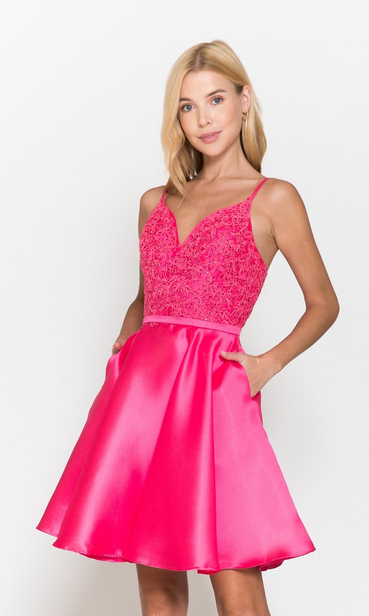 A woman models the Short A-Line Homecoming Dress with Pockets 8694 in fuchsia, featuring a lace bodice and satin skirt, standing with hands in pockets against a plain white background.