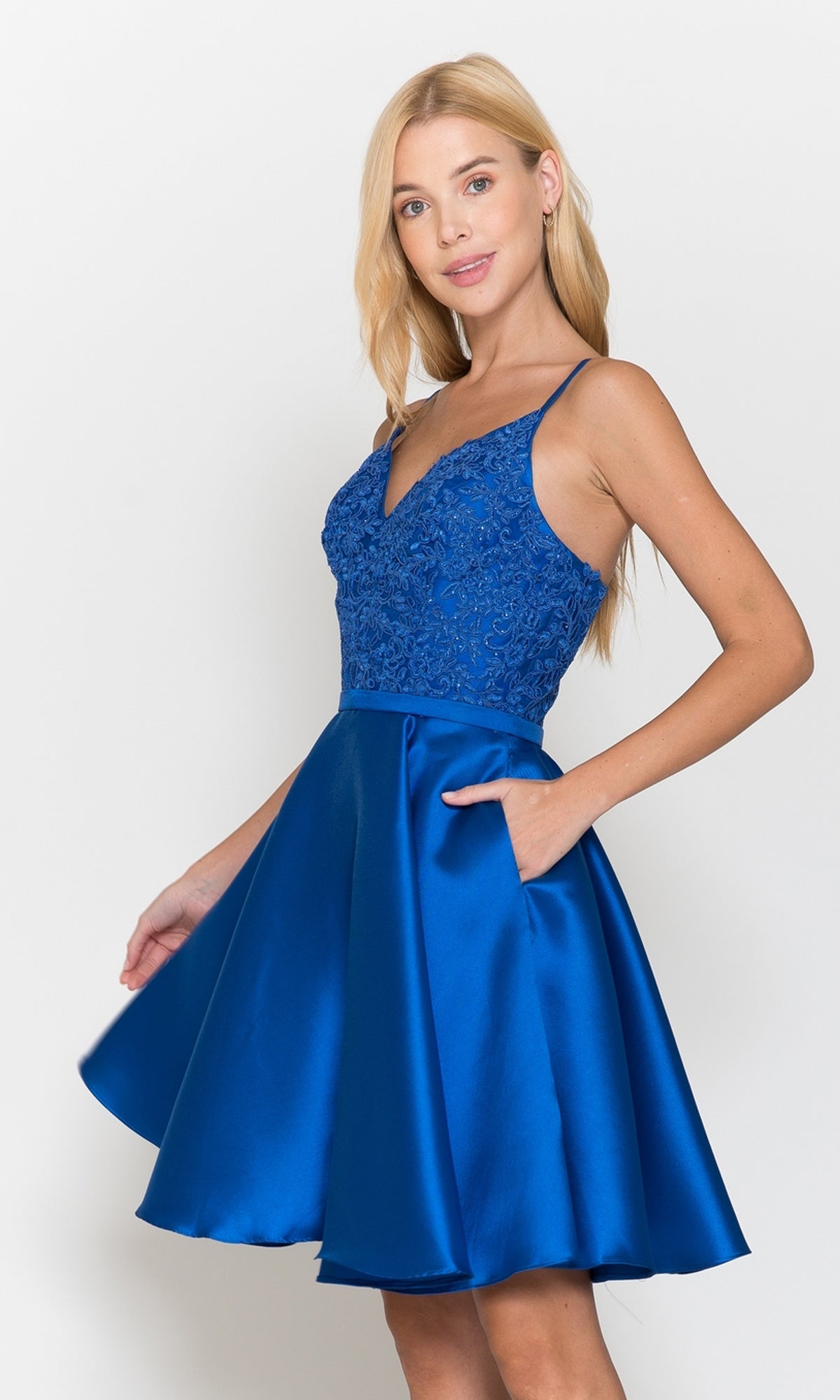 A woman models the Short A-Line Homecoming Dress with Pockets 8694 in royal blue, showcasing lace detailing on the bodice and a satin skirt, set against a plain white background.