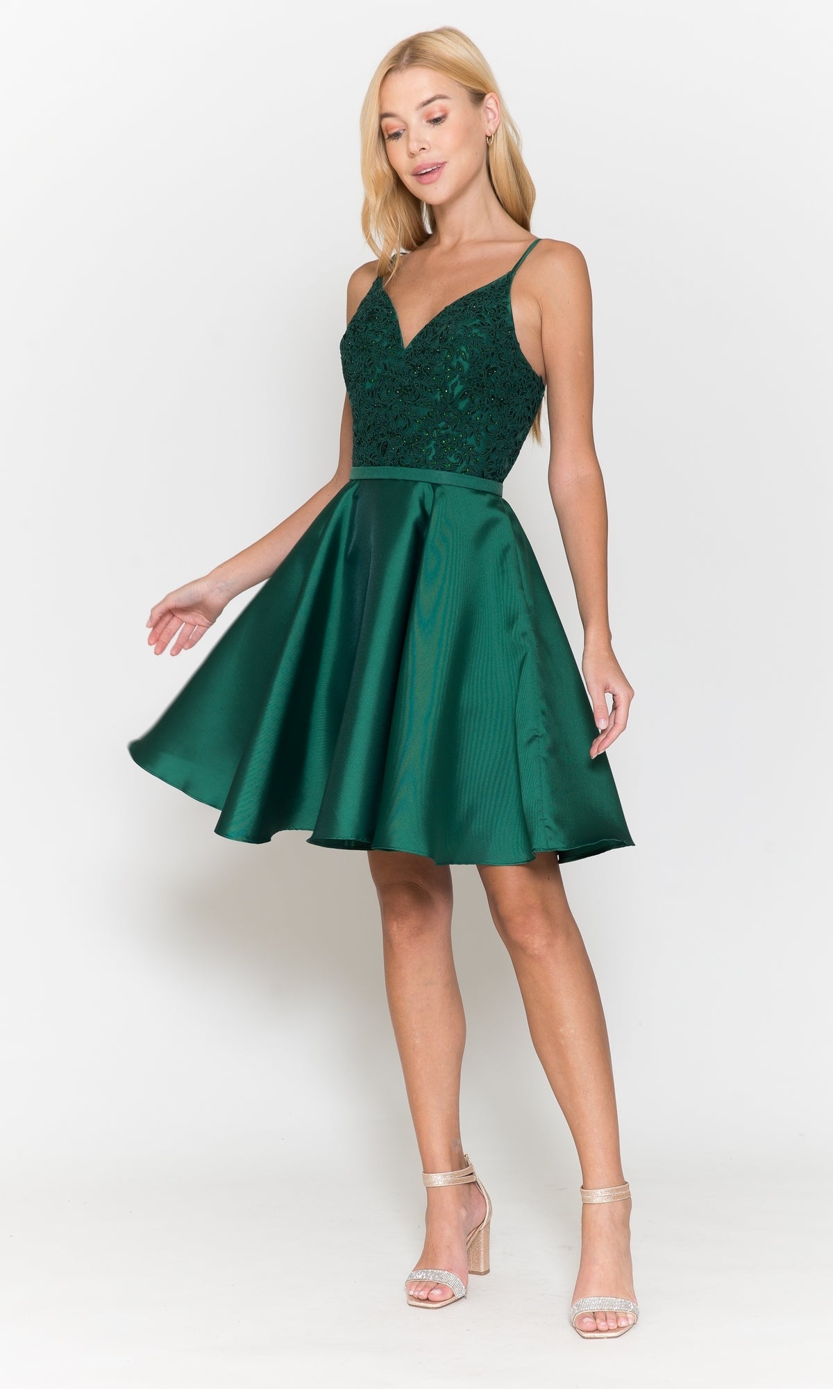 A woman wears the Short A-Line Homecoming Dress with Pockets 8694, featuring a sleeveless dark green lace bodice and flared satin skirt, paired with strappy beige heels, standing against a white background.