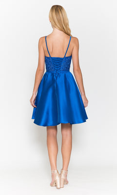 A woman faces away, wearing the Short A-Line Homecoming Dress with Pockets 8694 in royal blue, featuring a beaded V-neck bodice and lace-up corset back, paired with nude heels.