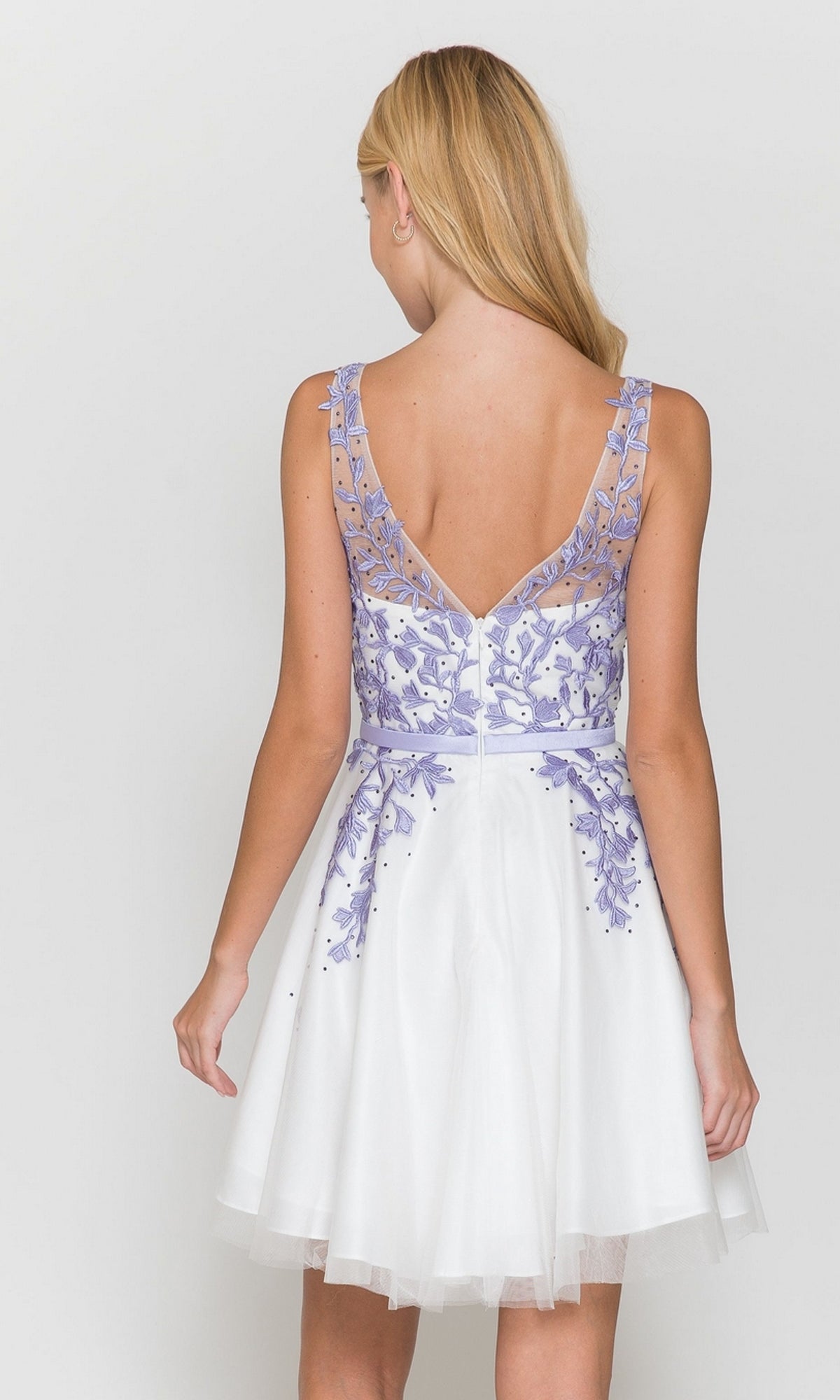 A woman with blonde hair models the Tulle and Lace Short A-Line Homecoming Dress 8696, showcasing its sleeveless white design with a sheer, lavender-embroidered bodice and flared skirt from the back against a plain background.