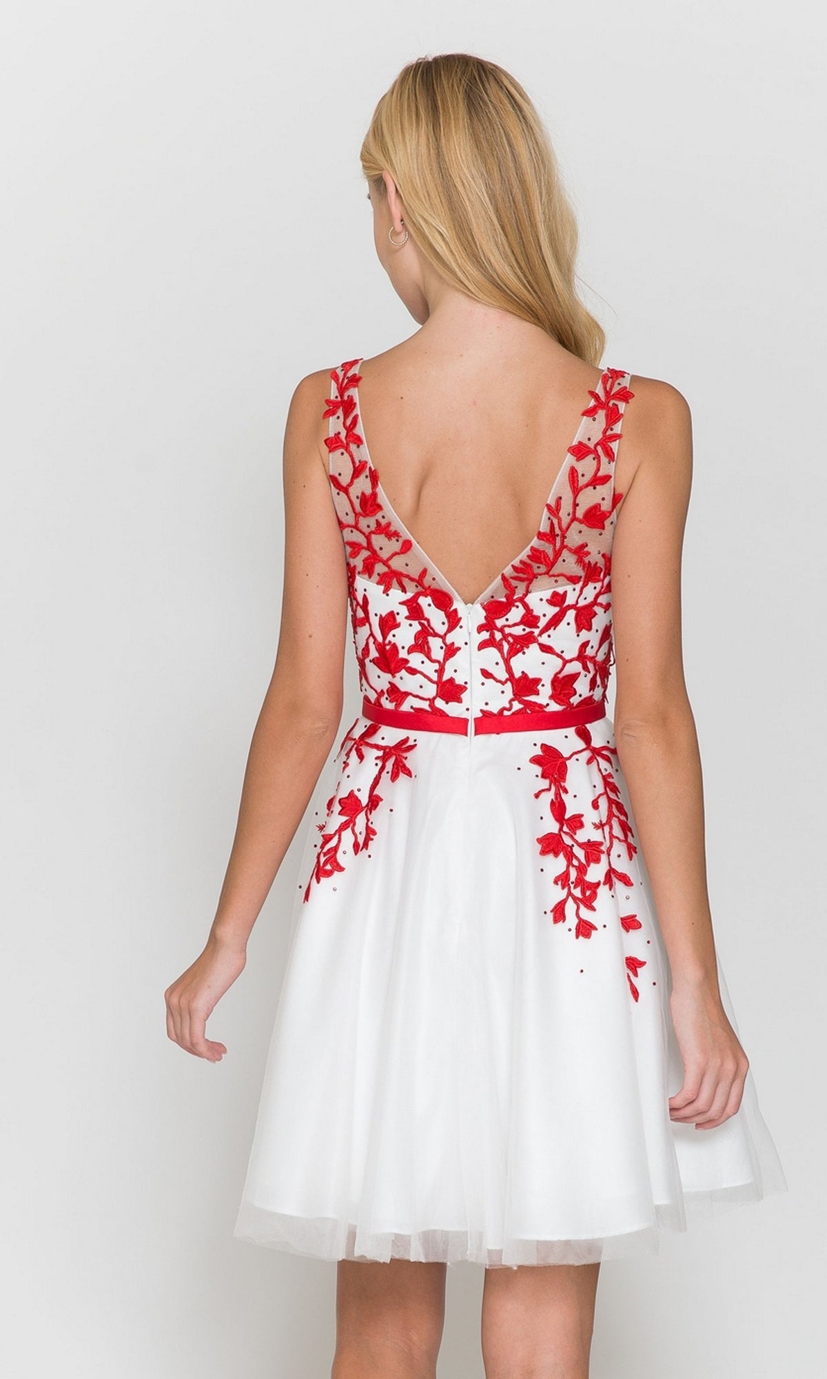 A woman faces away, wearing the Tulle and Lace Short A-Line Homecoming Dress 8696, featuring a sleeveless white design with a deep V-back and red floral embroidery on the bodice and waist.