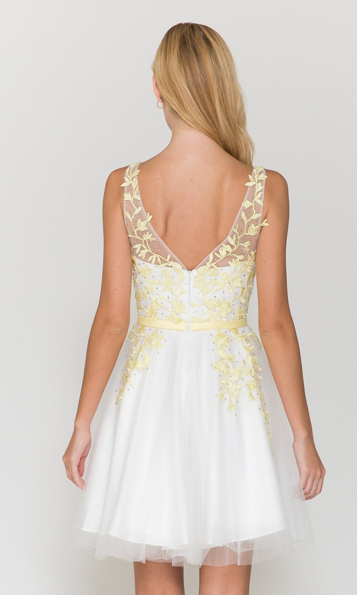 A woman with long blonde hair, seen from behind, wears the Tulle and Lace Short A-Line Homecoming Dress 8696—a sleeveless white dress with yellow floral embroidery and a sheer V-back.
