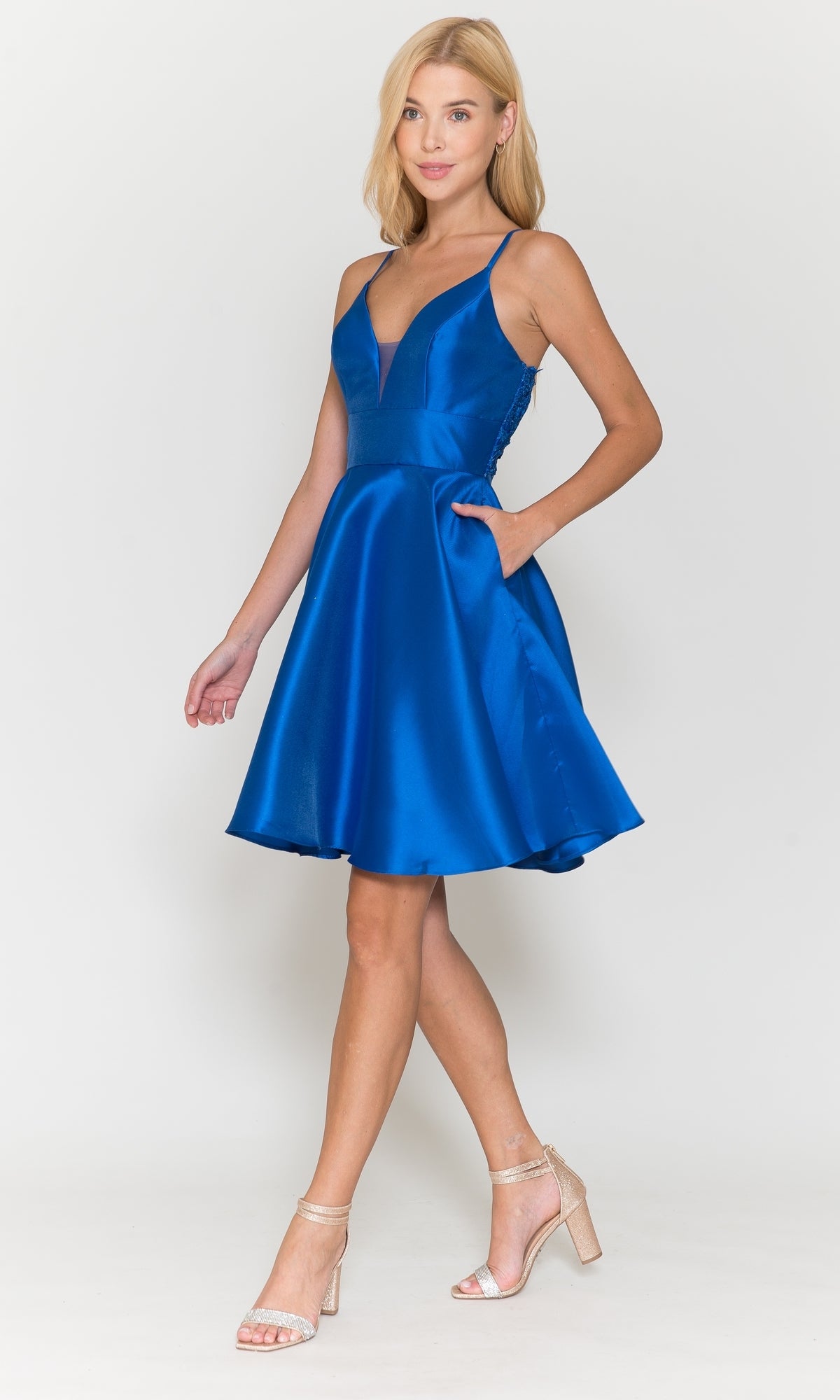 A woman wears the Lace-Back Short A-Line Homecoming Dress 8698, a sleeveless, knee-length royal blue dress with a V-neckline and side pockets, paired with nude high-heeled sandals, standing against a white background.