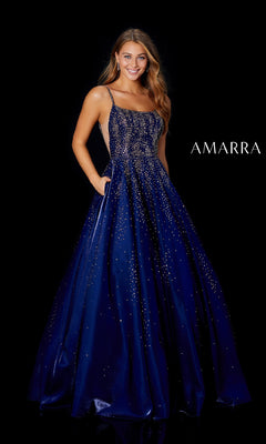 A woman in the Amarra 87292 long prom dress, featuring a navy blue color, thin straps, sparkling embellishments, and a satin A-line skirt, stands against a black background with "AMARRA" beside her.