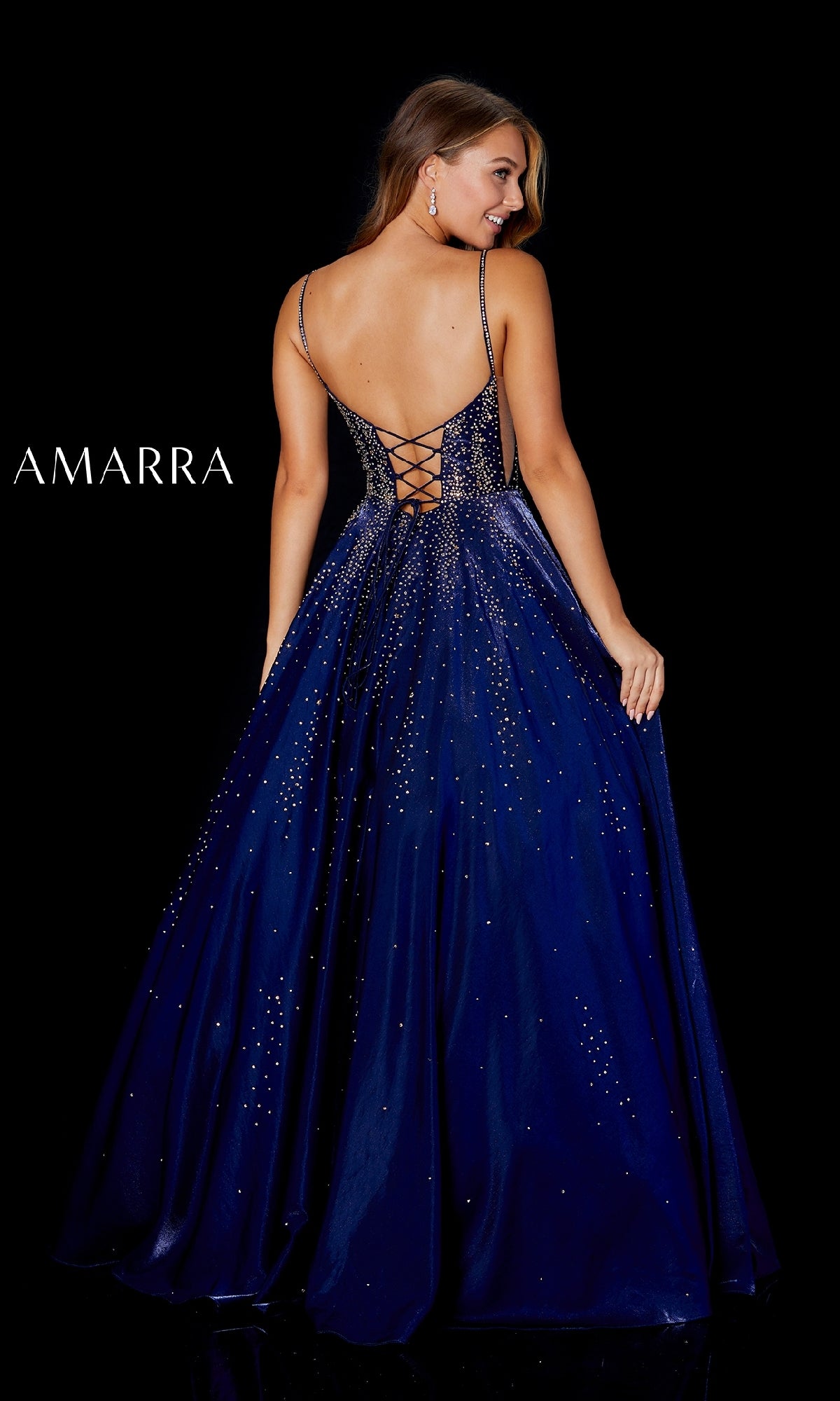A woman in the Amarra 87292 long prom dress with a sparkling deep blue design and lace-up back stands facing away against a black background. 