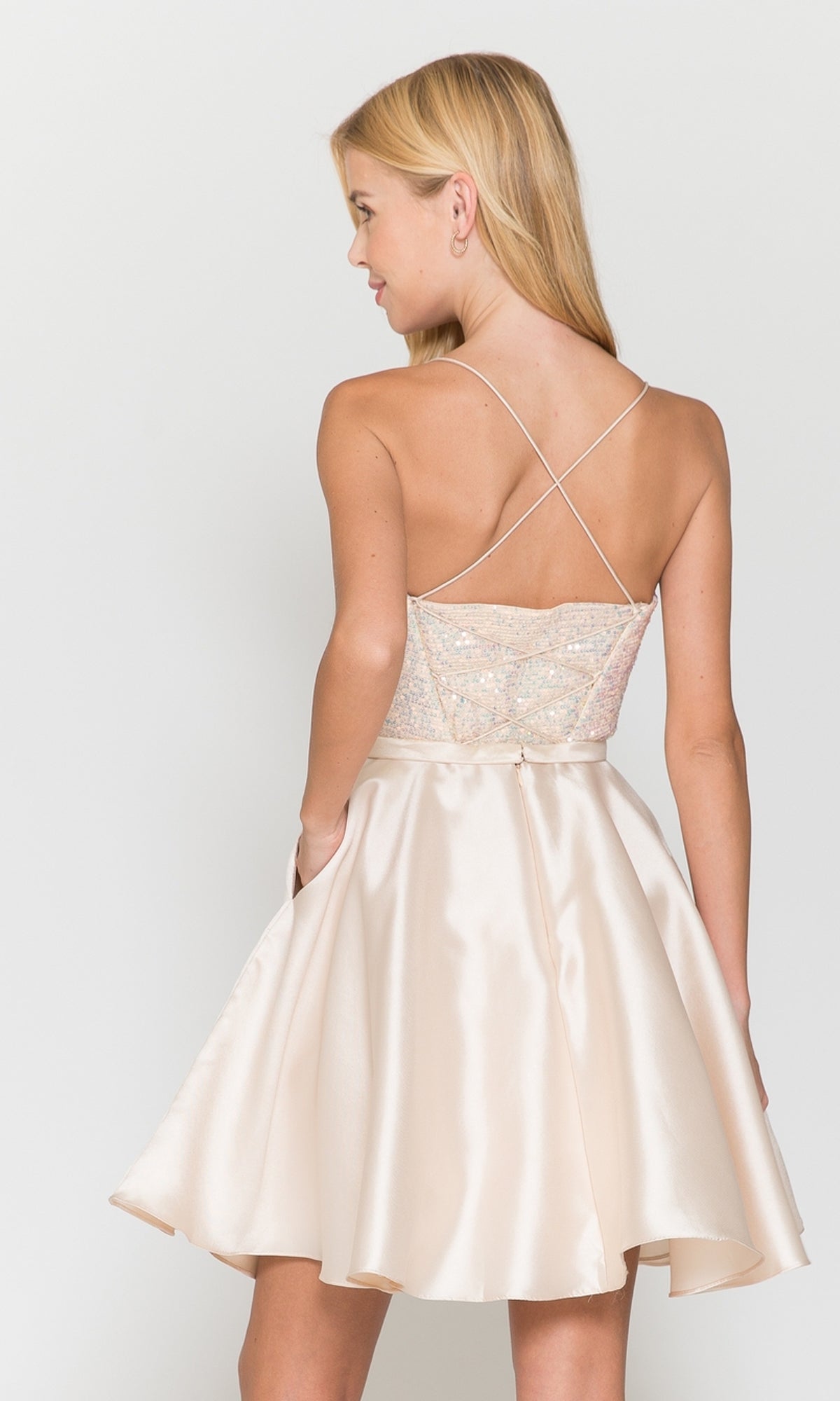 A woman with long blonde hair wears the Sequin-Bodice Short Flared Homecoming Dress 8730—a light pink, sleeveless A-line dress with a fitted, sequined bodice and flared satin skirt. She is facing backward, her head turned to the side.