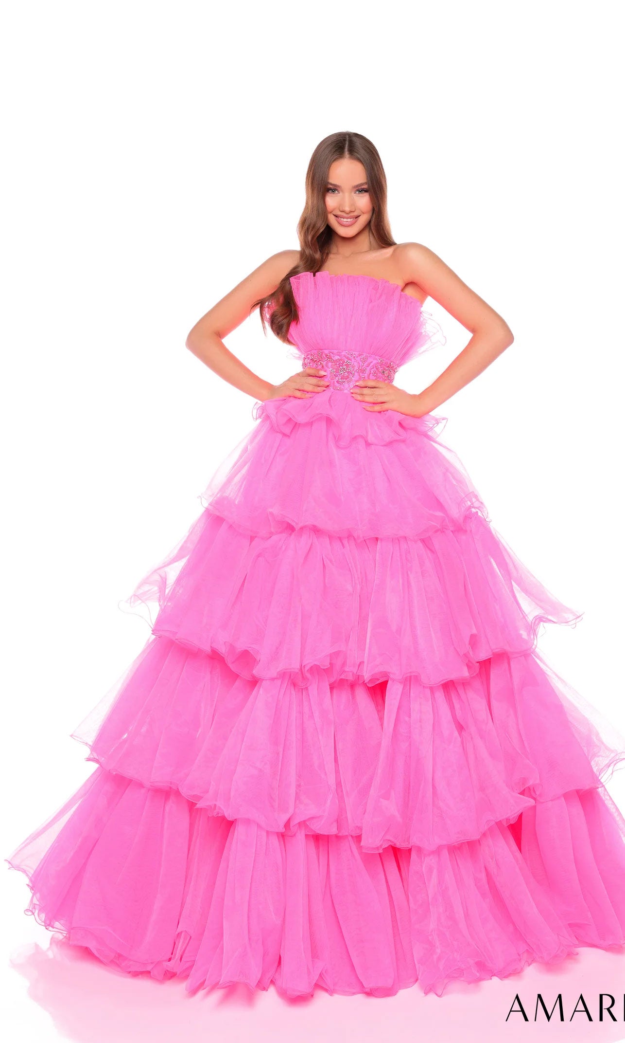 A woman smiles with her hands on her hips, wearing the Long Formal Dress 88083 by Amarra?a bright pink, strapless gown with a beaded waist?against a white background.