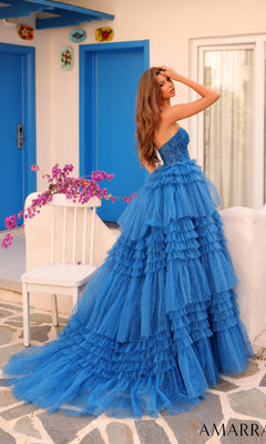 A woman wears the Long Formal Dress 88099 by Amarra, a strapless ball gown with a sheer corset bodice, standing on a stone patio near a white chair, in front of blue doors and decorative wall art.