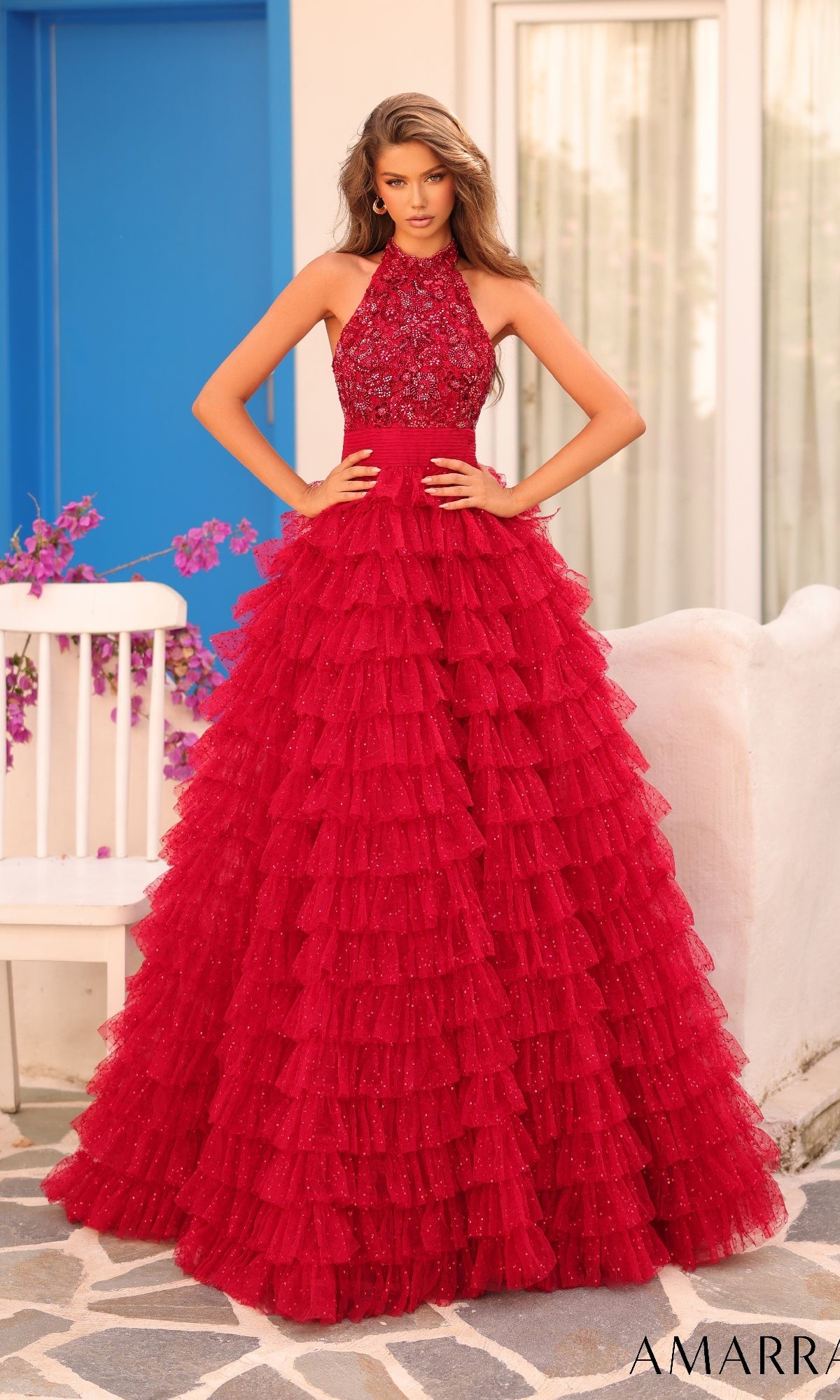 A woman stands outdoors near a white chair and blue door, wearing the Amarra Long Formal Dress 88102?a red A-line ball gown with a halter neck, beaded bodice, and ruffle layered skirt.