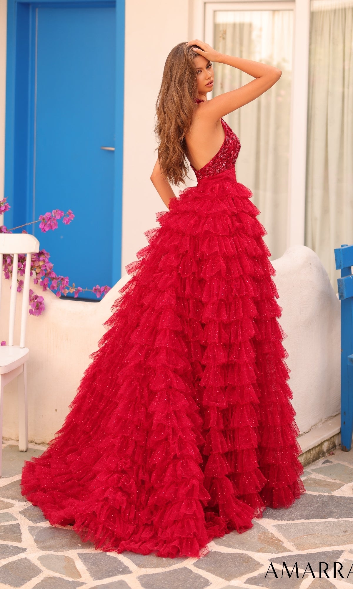 A woman in the Long Formal Dress 88102 by Amarra, a deep red ruffle layered gown, stands outdoors on a stone path near a white chair, blue door, and window, with blooming flowers in the background.