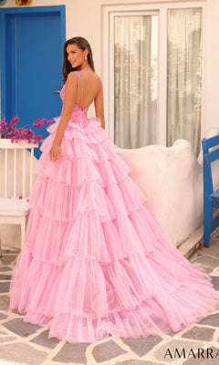 A woman wears the Long Formal Dress 88147 by Amarra, featuring a pink one-shoulder design with a ruffle-tiered overskirt, standing outdoors on a stone patio beside a white bench with flowers near blue doors and windows.