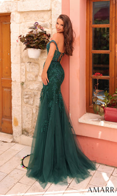 A woman in the Amarra 88224 long prom dress?an off-the-shoulder, fitted emerald green gown with a corset bodice and lace appliques?smiles over her shoulder outside near a window and potted plants.